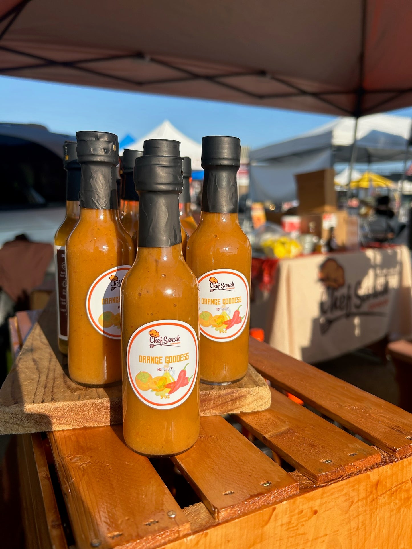 Bottles of orange goddess hot sauce with black caps on a wooden crate at an outdoor market.