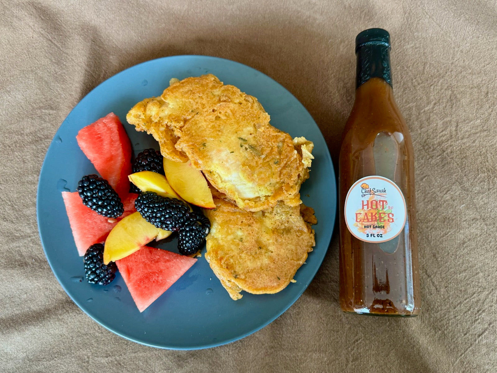 Blue plate with pancake-battered chicken and fruit, next to a bottle of hot sauce on a beige surface