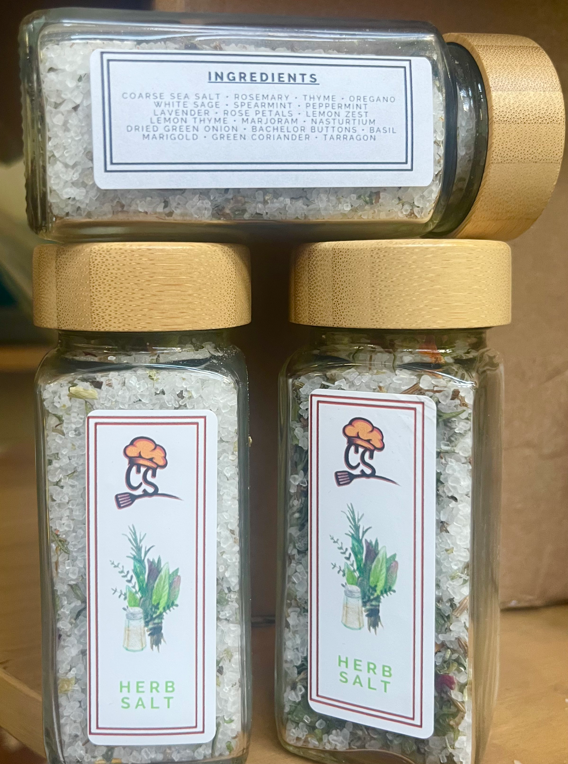 Two jars of herb salt with labels on a wooden surface. A third bottle shows the ingredients label.