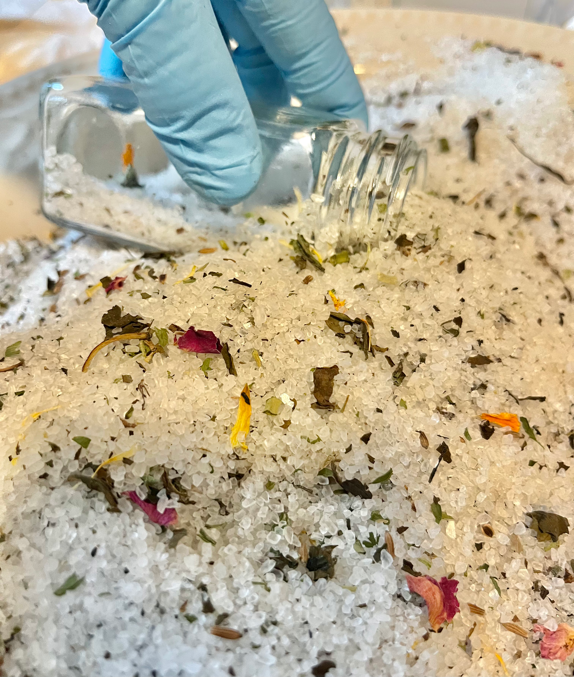 Hand with blue glove using a glass bottle to capture the salt mixture, herbs, and flower mixture.