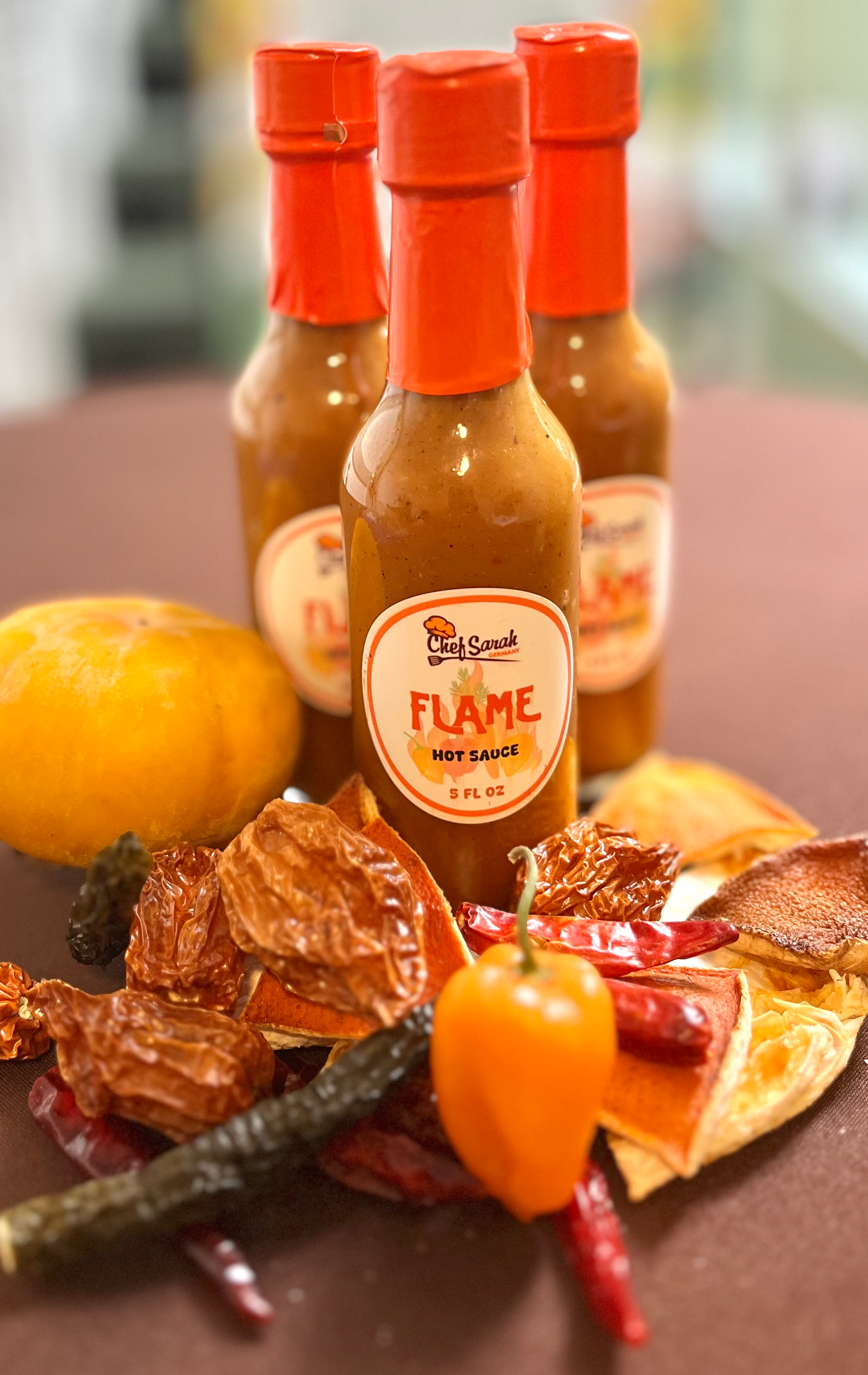 Three bottles of 'Flame' hot sauce on a table with peppers and a blurred background