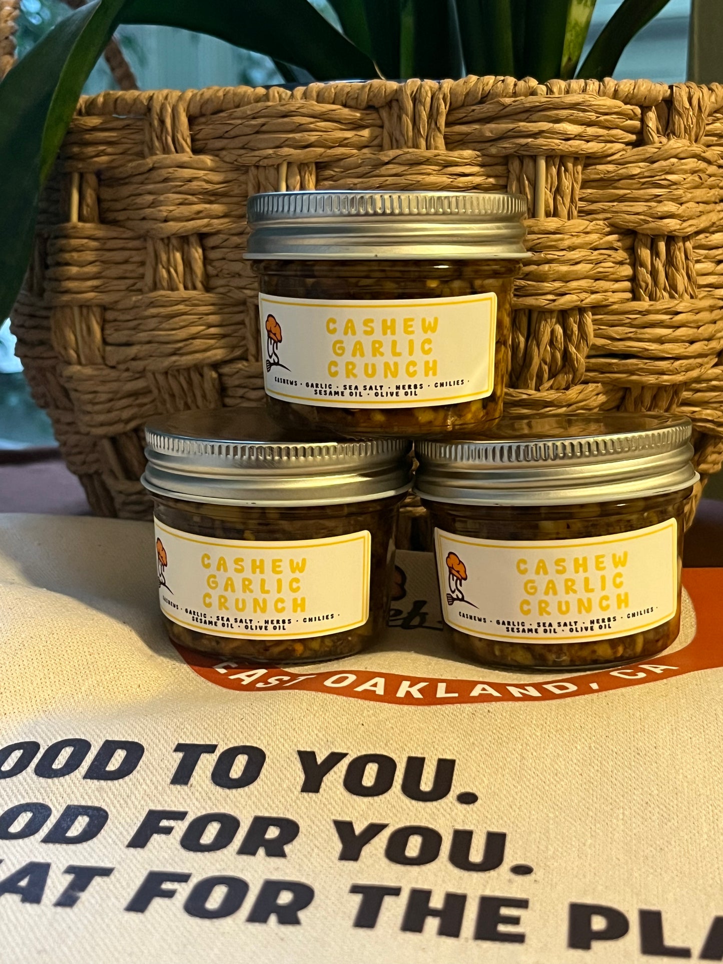 Three jars of 'Cashew Garlic Crunch' on a table with a woven basket in the background.