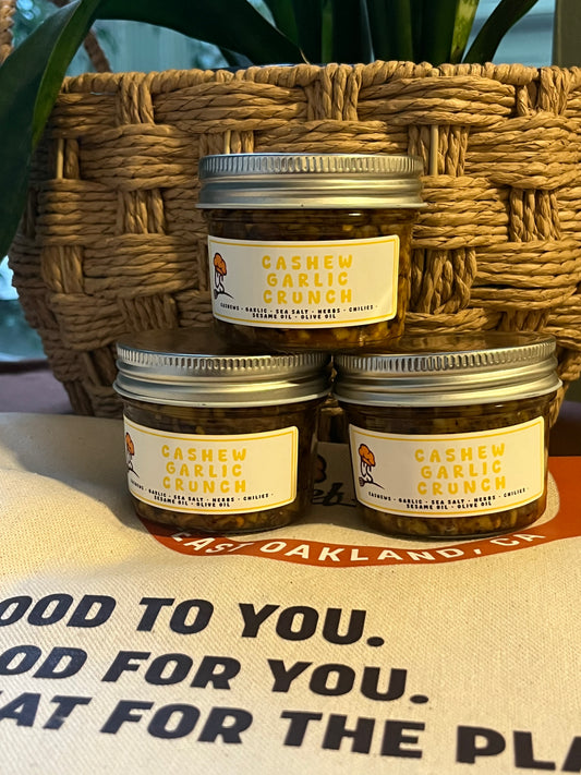 Three jars of 'Cashew Garlic Crunch' on a table with a woven basket in the background.