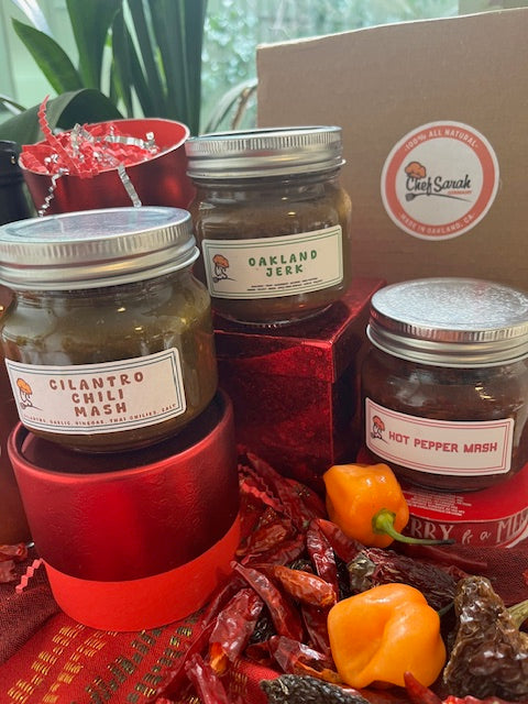 Jars of hot pepper jelly with labels on a red cloth, surrounded by peppers and a box.