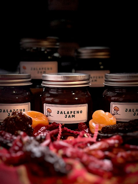 5 jars of jalapeno jam highlighted behind dried chilies on a red surface