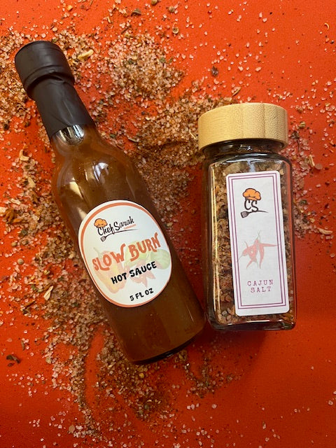 One bottle of 'Slow Burn' hot sauce and a jar of cajun salt on a red surface with visible spices.