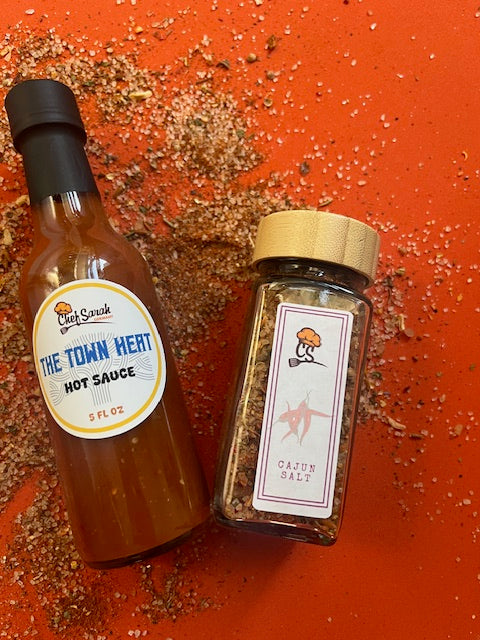One bottle of hot sauce and a jar of cajun salt on a red surface with visible spices.