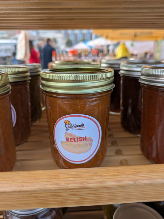 Jar of relish with a label on a wooden shelf at an outdoor market.