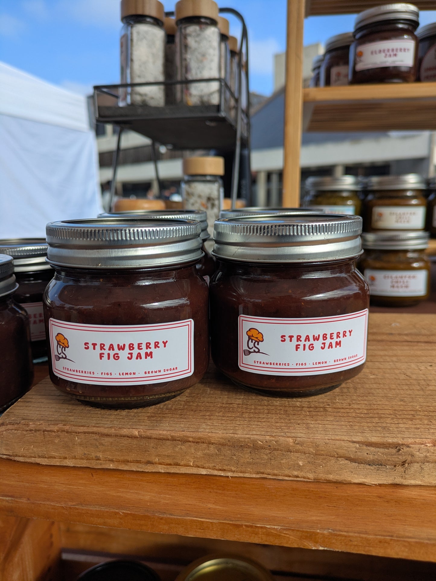 Two jars of strawberry fig jam on a wooden surface with a market background.