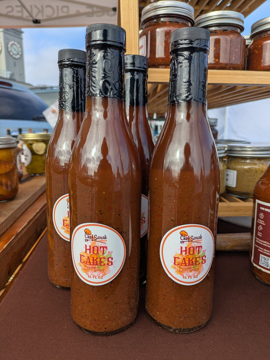 Bottles of 'Hot Cakes' hot sauce on a table with jars in the background.